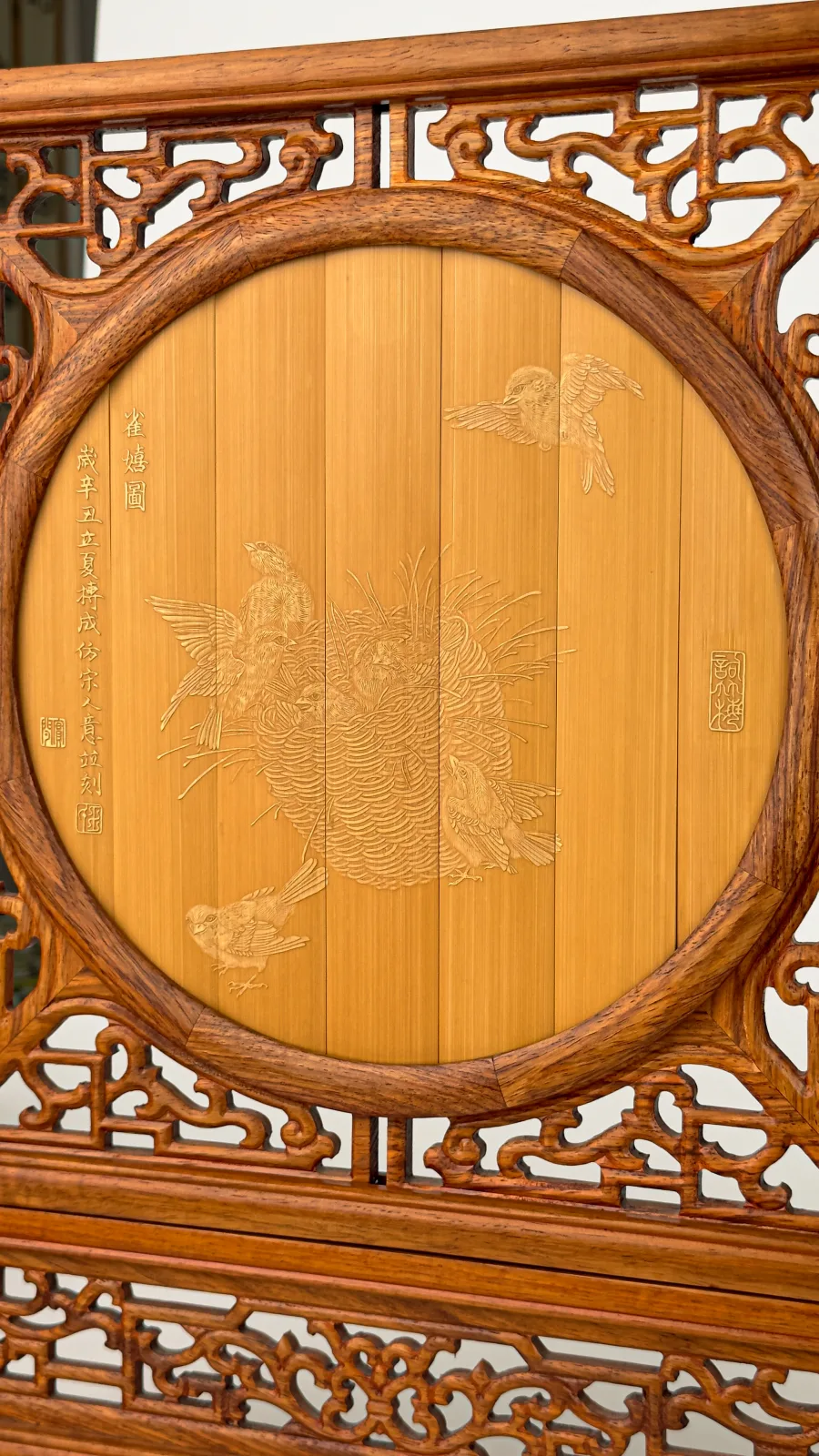 A round bamboo panel carved with sparrows around a nest, set in a carved hardwood screen, with a vertical inscription and small seals